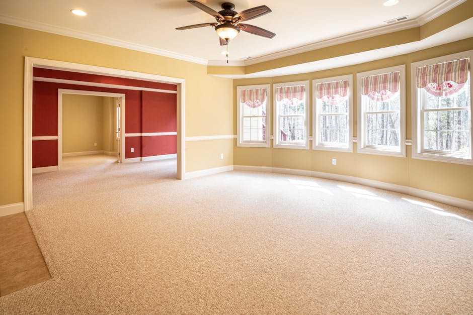An elderly man with grey hair and a beard is vacuuming a patterned area rug on a wooden floor of a bright, minimalist living room with large windows and natural light. He is wearing a light grey long-
