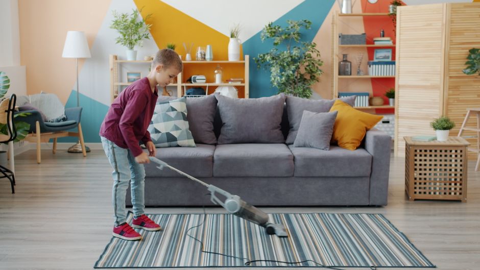 A young boy vacuuming a beige carpet in a bright living room with two sofas, one with a blue cushion and the other with an orange cushion, during daytime. An older man with gray hair, seated on one of