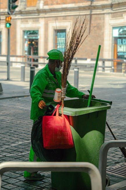 A street cleaner dressed in a bright green uniform and cap, wearing a protective face mask, using a broom and dustpan next to a large green waste bin on a paved urban sidewalk. The scene is illuminate