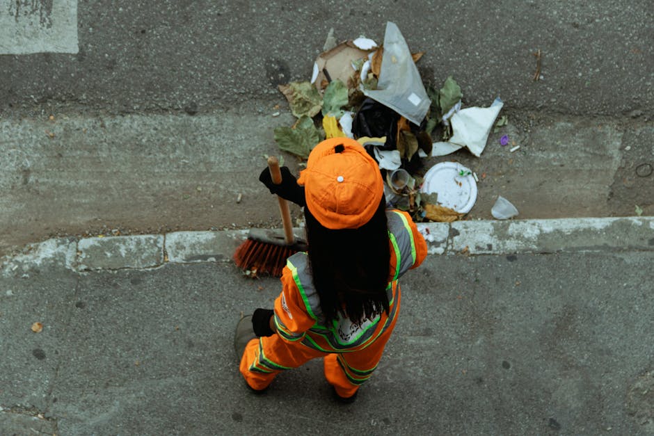 A street cleaner dressed in a bright green uniform and cap, wearing a protective face mask, using a broom and dustpan next to a large green waste bin on a paved urban sidewalk. The scene is illuminate